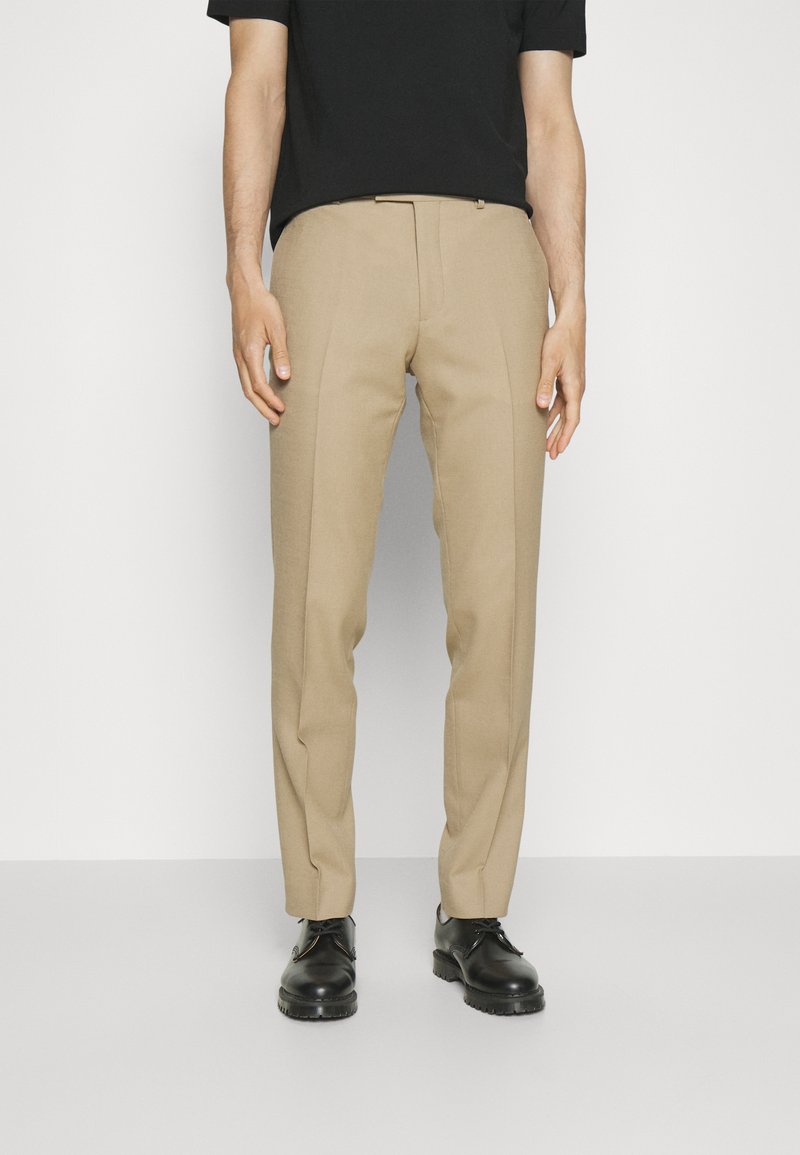 Beige, slim fit szabású nadrág, textúrált anyagból készült. Precíz elülső redők és standard derékrész jellemzi. Fekete cipőt visel.