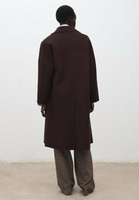Manteau en laine marron à coupe droite, avec des manches longues et une fente dans le dos. Associé à un pantalon à carreaux gris, vu de dos contre un mur clair.