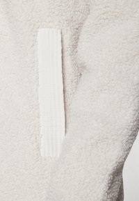 Fleece jakna v kremni barvi z teksturirano, mehko površino. Ima belo teksturirano tkanino kot dodatek na strani.