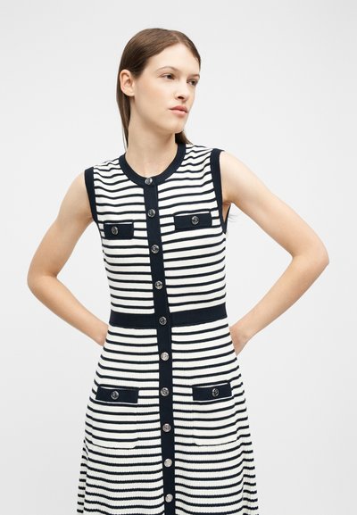 Femme aux cheveux bruns raides portant une robe sans manches à rayures horizontales noires et blanches avec des boutons et des poches, les mains sur les hanches.