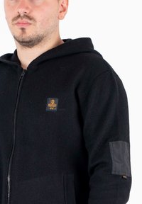 Felpa nera con zip in materiale felpato, dotata di una toppa in pelle sulla manica e una toppa con logo sul petto. Polsini e fondo a coste.