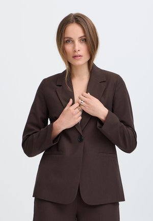 Jonge vrouw met lichtbruin haar, gekleed in een donkerbruin pakjasje en bijpassende broek, die het jasje aan de voorkant rechtzet tegen een effen lichte achtergrond.