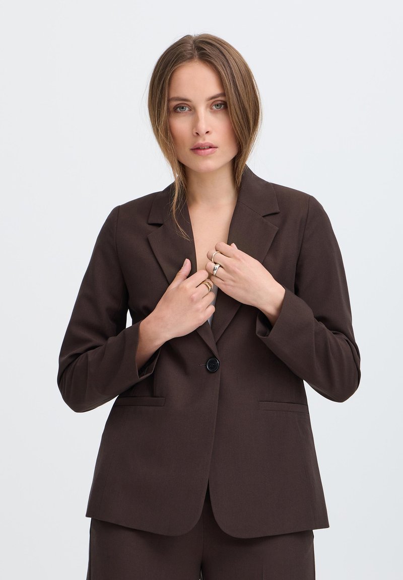 Jeune femme aux cheveux châtain clair portant une veste de costume marron foncé et un pantalon assorti, ajustant le devant de la veste sur un fond clair uni.