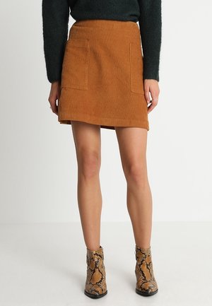 Vrouw draagt een bruine ribfluwelen minirok met steekzakken aan de voorkant, een zwart shirt met lange mouwen en enkellaarsjes met slangenprint, staand tegen een witte achtergrond.