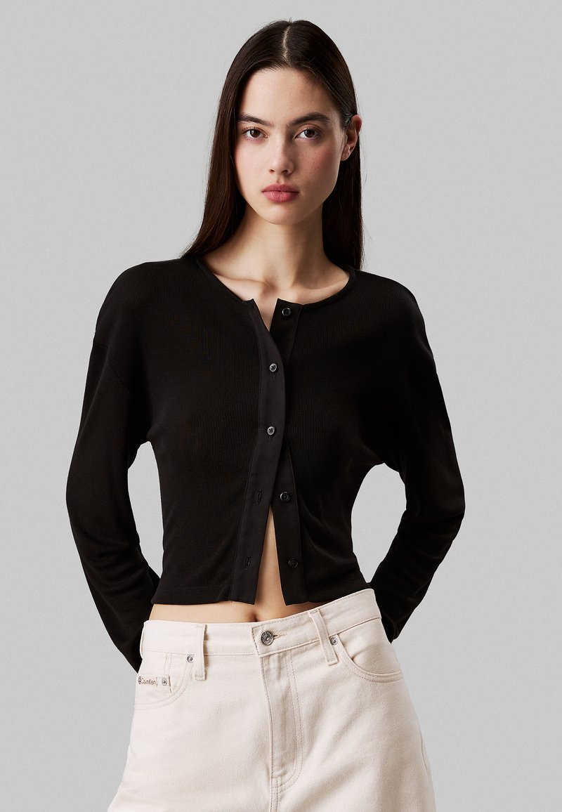 Cardigan lungo a costine nera, con scollo rotondo e chiusura a bottoni. Abbinato a jeans chiari, che mostrano un design cropped.