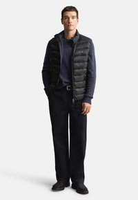 Schwarze, gesteppte Weste mit hohem Kragen, getragen über einem dunkelblauen Pullover. Kombiniert mit dunkler Hose und schwarzem Schuhwerk, mit schlichtem Design.