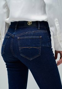 Vaqueros de mezclilla azul oscuro con un diseño ajustado. Incluye detalles como herrajes dorados, costuras en contraste y una etiqueta de cuero en la cintura.