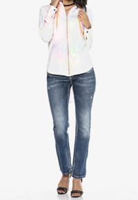 Chemise zippée blanche avec un éclat arc-en-ciel, cordons orange, associée à un jean en denim bleu avec des détails effilochés. Sandales à talons noires.