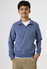 Sudadera azul de media cremallera con una textura suave, puños y dobladillo acanalados, y un pequeño detalle de logo en el pecho. Llevada sobre una camiseta blanca.