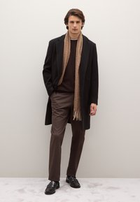 Abrigo negro, suéter de punto marrón, pantalones marrones y una bufanda de textura color tostado. Mocasines negros completan el conjunto. Fondo neutro.