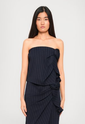 Young woman with long dark hair wearing a strapless navy pinstripe dress featuring layered, ruched floral fabric details.