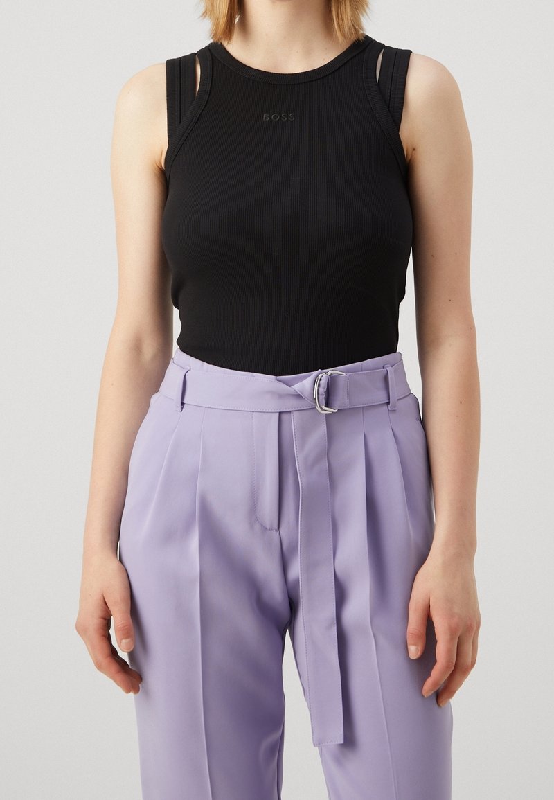 Femme portant un haut noir sans manches à fines bretelles et un pantalon lavande taille haute avec une ceinture assortie sur fond uni.