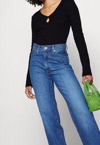 Schwarzes Langarmoberteil mit einem Schlüssellochdetail und hoch taillierten blauen Denim-Jeans. Handgehaltene grüne Perlenhandtasche sorgt für Textur und Farbkontrast.