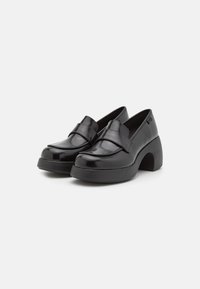 Chaussures en cuir verni noir avec une finition brillante, présentant une large sangle sur le cou-de-pied et un talon large et courbé.
