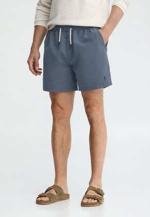 Man wearing blue drawstring shorts and brown buckle sandals, standing with one hand in pocket against white background.