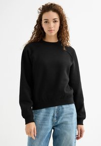Sudadera de algodón negra con cuello redondo, puños y dobladillo de canalé, combinada con jeans de color azul claro. Presenta un ajuste relajado y mangas raglán.