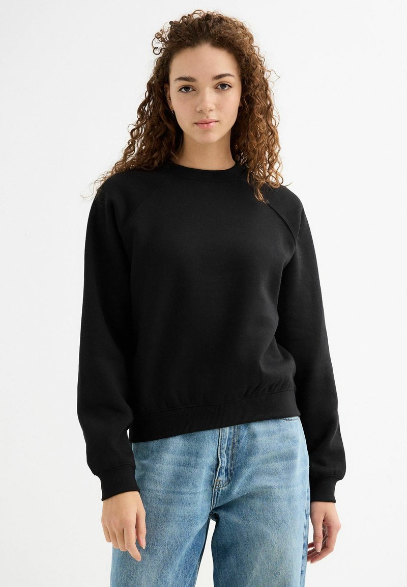 Sudadera de algodón negra con cuello redondo, puños y dobladillo de canalé, combinada con jeans de color azul claro. Presenta un ajuste relajado y mangas raglán.