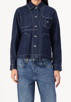 Dunkelblaue Jeansjacke mit Knöpfen, Kontrastnähten und Brusttasche mit Klappe, kombiniert mit hellblauen Jeans.