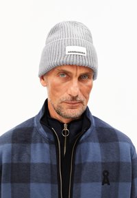 Grijze geribde gebreide beanie met een wit label met "ARMEDANGELS." Het model draagt een blauwe ruitjes zip-up jas met zwarte accenten.