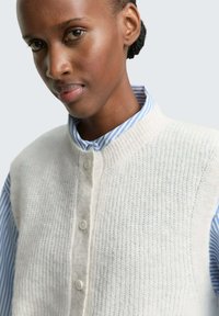 Cardigan sans manches en tricot blanc avec un motif texturé, présentant un col arrondi et une fermeture à boutons, porté sur une chemise à rayures bleues.