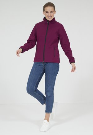 Maroon softshell jacket with a high collar, zip front, and two zippered side pockets. Worn with fitted blue jeans and white sneakers.