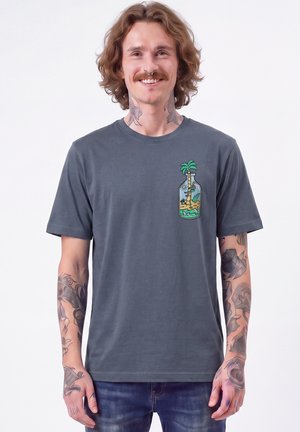 Lächelnder Mann mit lockigem Haar und Tattoos trägt ein graues T-Shirt mit einer Flasche, die eine Strandszene und eine Palme auf der Brust zeigt.