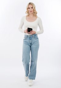 Mujer rubia con top blanco de manga larga y jeans azul claro sostiene una cartera negra, de pie frente a un fondo blanco.