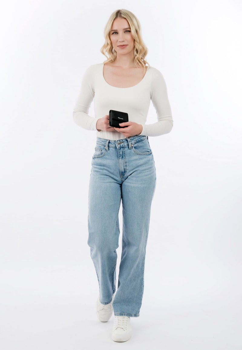 Mujer rubia con top blanco de manga larga y jeans azul claro sostiene una cartera negra, de pie frente a un fondo blanco.