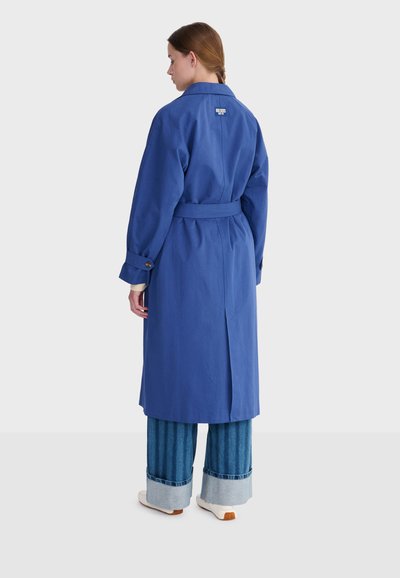 Femme vue de dos portant un long manteau bleu ceinturé, un jean large avec des revers et des chaussures blanches sur un fond gris uni.