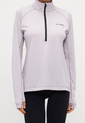 Femme portant un pull violet clair à manches longues avec fermeture éclair à mi-hauteur et logo Columbia Titanium, ainsi que des leggings noirs, debout devant un fond uni.