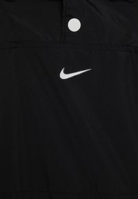 Sort Nike-jakke med hvidt logo, glat tekstur og knapdetalje ved kraven, designet til et strømlinet udseende.