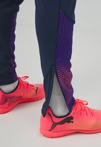 Hosen in Marineblau mit lila gemusterten Seitenteilen, ausgestattet mit Reißverschlüssen. Helle pinke Sportschuhe mit einem Farbverlauf und schwarzen Logoakzenten.