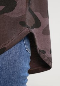 Brun topp med camouflasjemønster og buet nederkant, laget av myk stoff, kombinert med blå jeans som har en synlig baklomme og lyse sømmer.