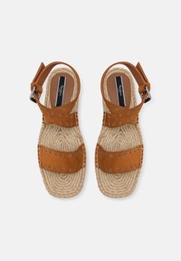 Paire de sandales en daim marron avec brides de cheville et œillets métalliques, semelles en corde tissée et formes d'orteils rectangulaires sur fond blanc.
