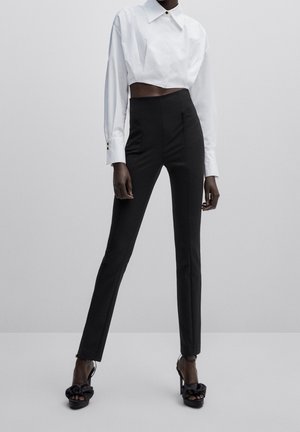 Person, die ein weißes, kurzes Langarmshirt, hoch taillierte schwarze schmale Hose und schwarze Sandalen mit Absätzen und Schleifen trägt.