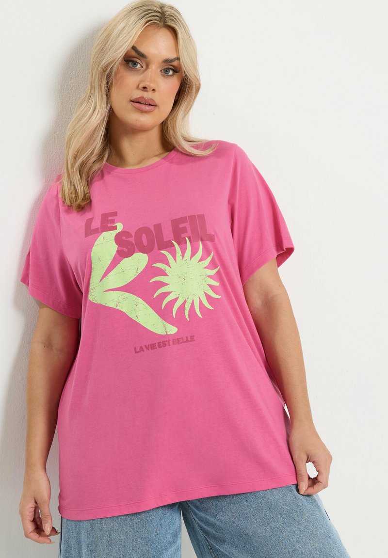 Blonde Frau in einem pinken, übergroßen T-Shirt mit grünem abstraktem Sonnen- und Blätterdesign sowie französischem Text, kombiniert mit hellblauen Jeans, vor einer weißen Wand.