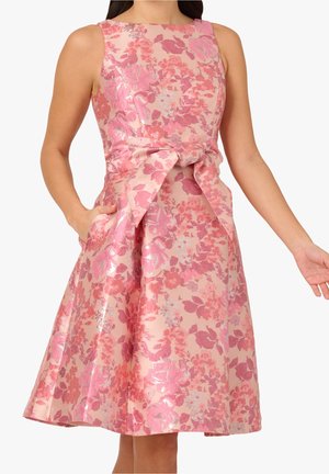 Różowa sukienka w kwiaty bez rękawów z metalicznym połyskiem, dopasowanym gorsetem, rozkloszowanym dołem i dużą kokardą z przodu, noszona przez kobietę z jedną ręką w kieszeni.