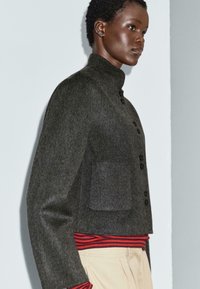 Vista laterale di una persona che indossa una giacca grigio scuro con bottoni sopra una camicia a righe rosse e pantaloni beige, su uno sfondo chiaro e semplice.