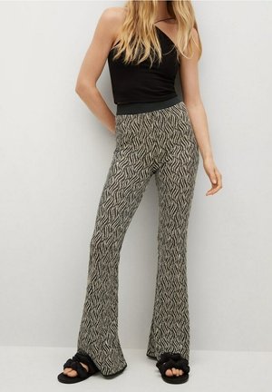 Kobieta ubrana w czarny top bez rękawów, spodnie z szerokimi nogawkami o wysokim stanie z czarno-białym abstrakcyjnym wzorem oraz czarne plecione sandały, stojąca na tle szarej ściany.