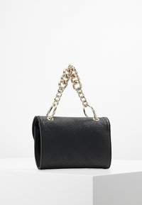 Musta tikattu käsilaukku, valmistettu teksturoidusta synteettisestä materiaalista, jossa on kullanvärinen ketjukahva ja pintaan painettu logodetalji.