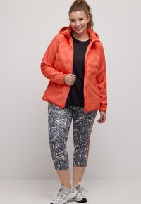 Oranžová péřová bunda s kapucí, v kombinaci se šedými mramorovanými legínami 3/4 s červenými bočními pruhy. Žena nosí šedé tenisky.