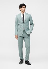 Άνδρας με σγουρά μαλλιά που φορά ανοιχτοπράσινο κοστούμι, λευκό πουκάμισο, μαύρη γραβάτα και μαύρα επίσημα παπούτσια, στέκεται με το ένα χέρι στην τσέπη.