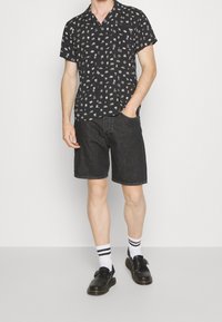 Maglietta nera a maniche corte con motivi bianchi, abbinata a pantaloni corti in denim scuro, calze bianche con strisce nere e scarpe nere.