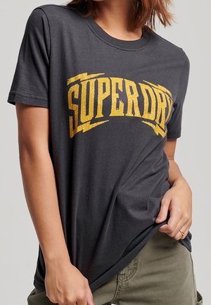 Osoba nosząca ciemnoszarą koszulkę z grubym żółtym napisem „SUPERDRY”, lekko podciągającą koszulkę w talii na oliwkowych spodniach.