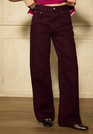 Persona che indossa pantaloni a gamba larga a vita alta di colore bordeaux scuro e scarpe nere con punta tonda, in piedi su un pavimento di legno con le mani che sistemano la vita.