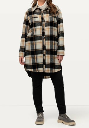 Woman wearing beige, black, and gray plaid long shirt jacket with black pants and gray lace-up ankle boots, standing against a plain background.