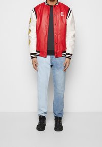 Röd lädervarsityjacka med vita ärmar, svarta ränder och logotyp. Kombinerad med ljusblå denimjeans och svarta sneakers.