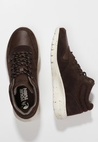 Paire de baskets en cuir marron foncé et suède avec des semelles blanches tachetées, des lacets noirs à motifs et des colliers de cheville rembourrés.
