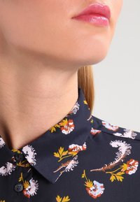 Granatowa bluzka w kwiaty z klasycznym kołnierzykiem, zapięciem na guziki i żywymi wzorami w kolorach pomarańczowym, białym i zielonym. Gładka tekstura materiału.