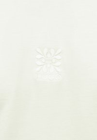 Λευκό ύφασμα με ανάγλυφο ανθικό σχέδιο πάνω από τη λέξη "HUGO" με ανάγλυφα γράμματα στο κέντρο.